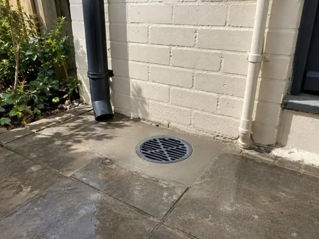 Outdoor drain grate set in paved courtyard beside a painted brick wall in Dulwich Hill