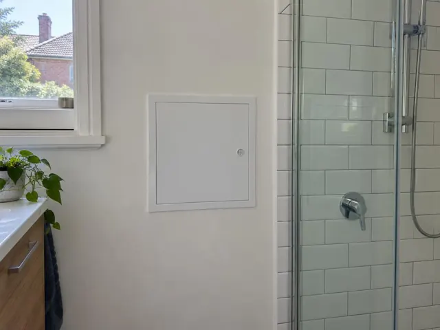 White access panel in a painted bathroom wall beside a tiled shower and window in Lewisham